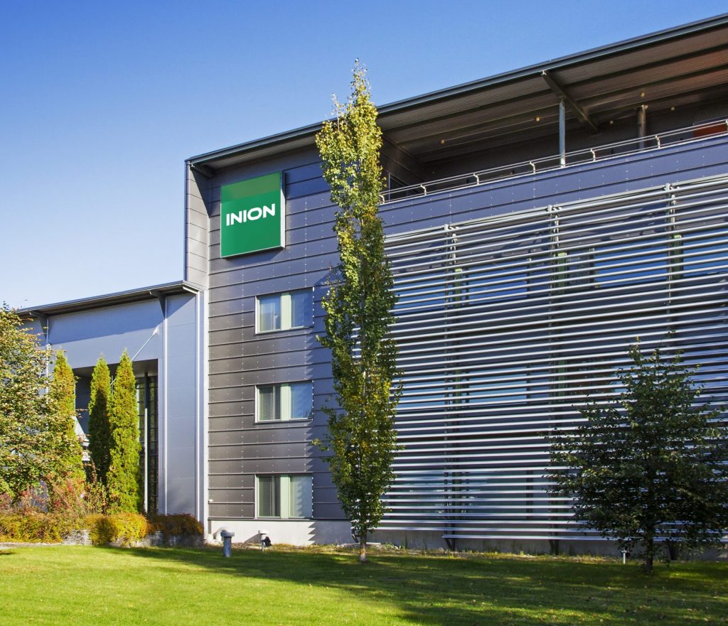 Inion headquarters building in Tampere Finland on a sunny day. The gray building has a green Inion logo close to the roof. Green trees and grass around the building.