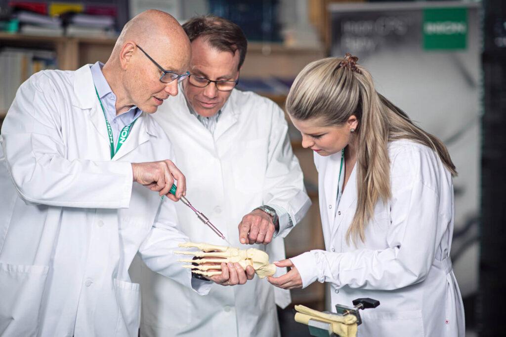 Team of experts standing close to each other wearing white doctor's jackets. One person is showing the other two where to install the bioabsorbable screw in a sawbone ankle model.